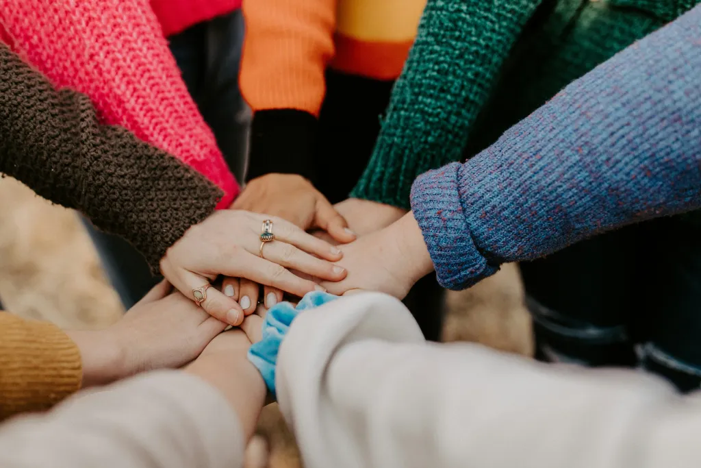 A diverse group of hands layered on top of each other, showcasing various skin tones and rings, with colourful knitted jumpers visible.