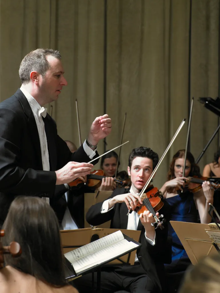 A conductor leading an orchestra, with musicians playing violins in formal attire, in a concert setting.