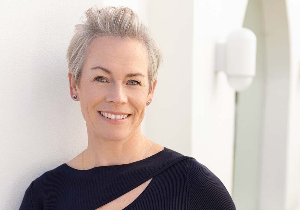 Portrait of Bronwen Downie, an executive coach and consultant, smiling while wearing a black top, with a modern architectural background.