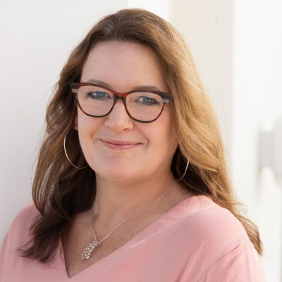 A woman with long brown hair, wearing glasses and a pink blouse, smiling softly against a light-colored background.