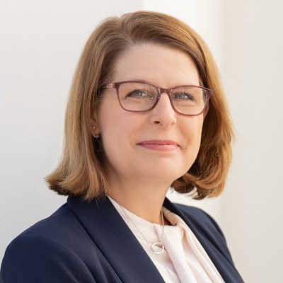 Professional headshot of a woman with shoulder-length brown hair, wearing glasses, and dressed in a dark blazer and light top, smiling pleasantly against a light background.