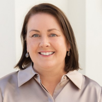 A woman with shoulder-length brown hair, wearing a light-colored blouse and earrings, smiling at the camera.