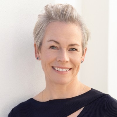 Smiling woman with short, styled gray hair, wearing a dark top, posing against a light background.