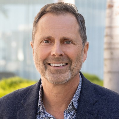 Smiling middle-aged man in a dark blazer and patterned shirt, standing outdoors with greenery and blurred background.