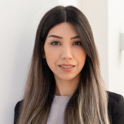 A professional portrait of a woman with long, straight, dark hair. She is wearing a light sweater and a blazer, standing against a neutral background.