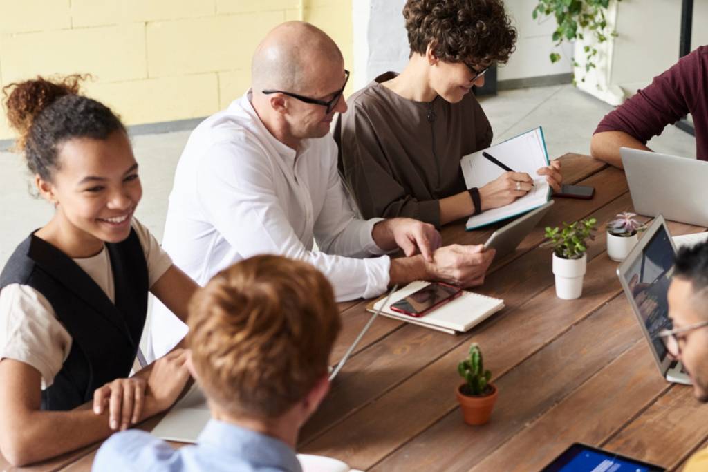 A diverse group of six people gathered around a wooden table in a modern workspace, engaged in discussions and collaborating on laptops and tablets. One person is writing in a notebook, while others are smiling and interacting, with small potted plants on the table.