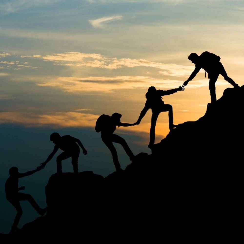 Silhouettes of hikers helping each other climb a rocky mountain at sunset.