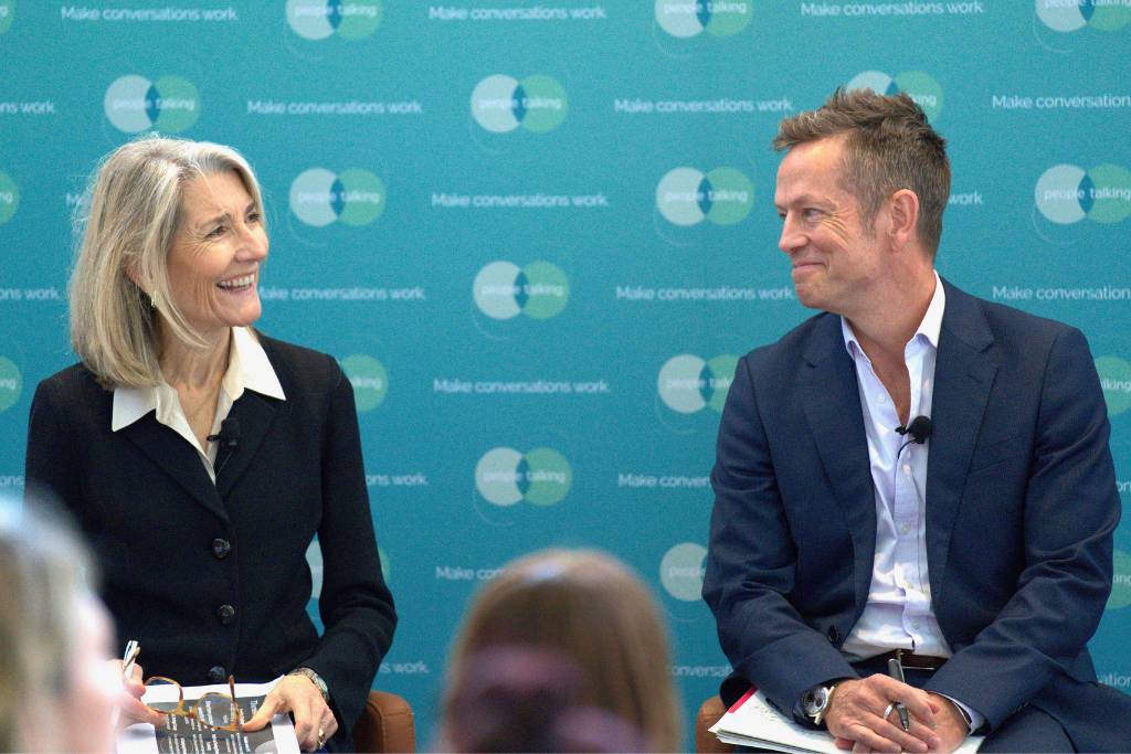 Two presenters engaging in a conversation on stage, smiling at each other, with a backdrop featuring the phrase 'Make conversations work'.