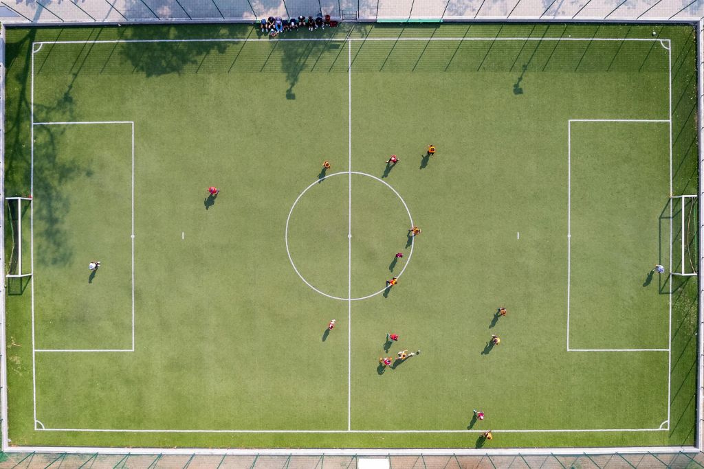 An overhead view of a soccer field with players in red and orange jerseys engaged in a match, surrounded by a green synthetic surface and goalposts.