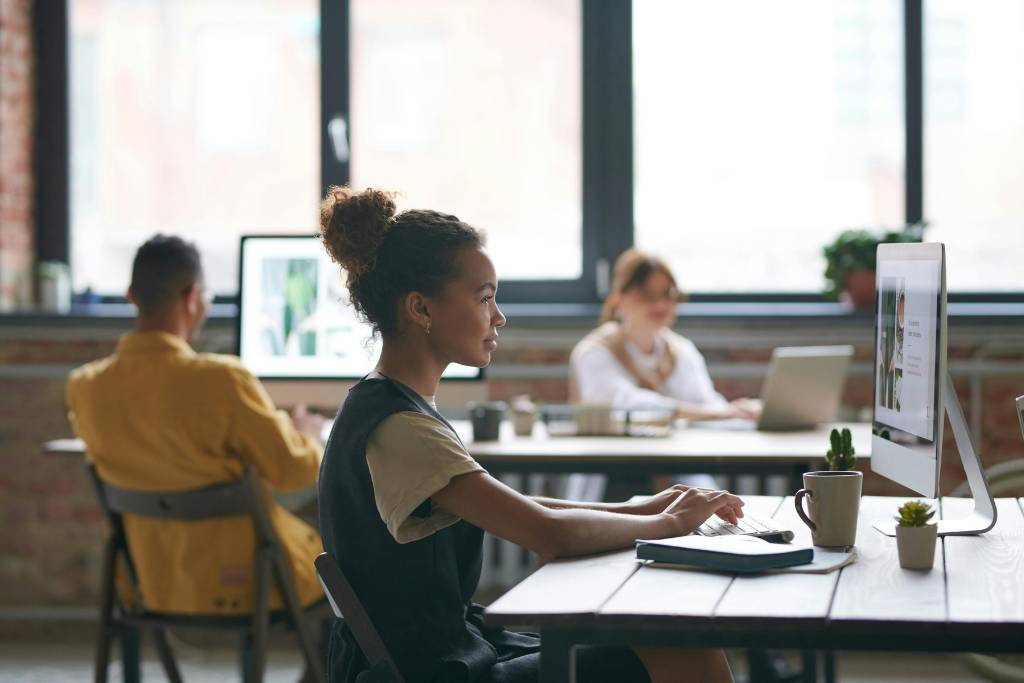 A young woman working on a computer in a modern office space, with two colleagues engaged in work at nearby desks.