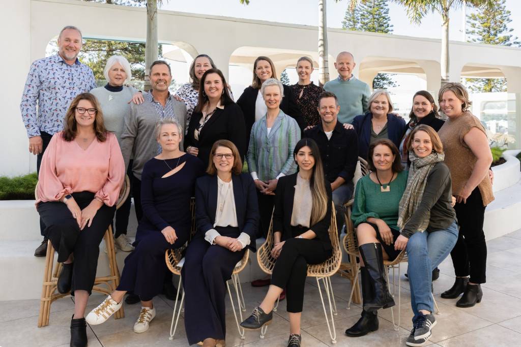 Group photo of a diverse team of 21 people standing and sitting together in an outdoor setting, featuring modern architecture and greenery in the background.