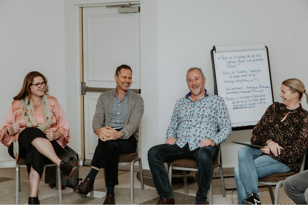 A group of four people seated in a semi-circle, engaged in an interactive discussion. They are in a bright room with a whiteboard displaying notes behind them, and appear to be sharing ideas and experiences.