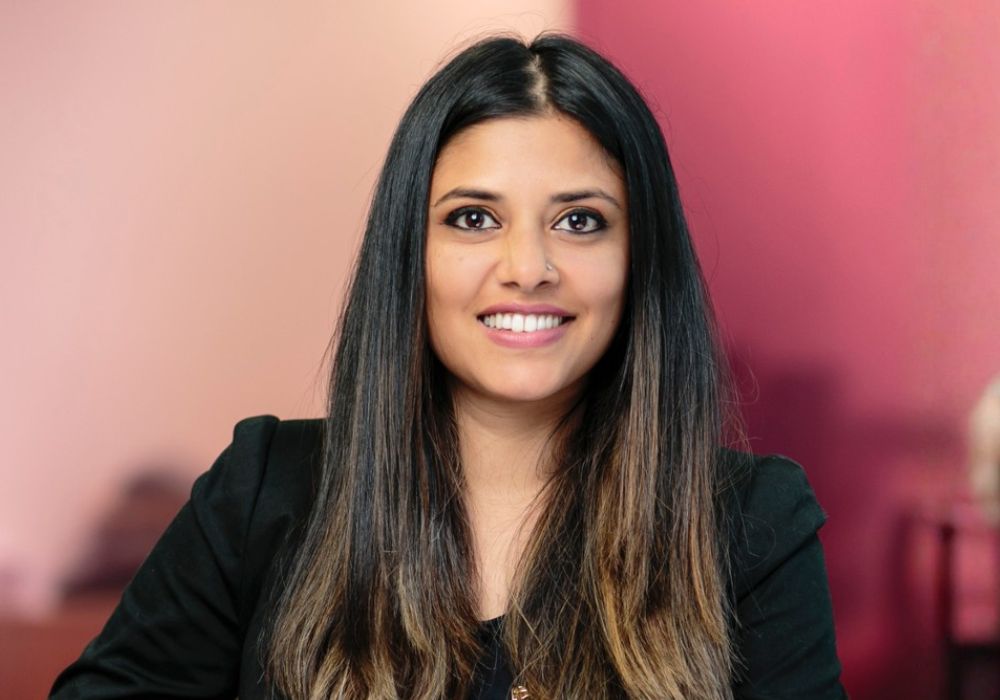 A woman with long, straight hair smiling at the camera, wearing a black blazer, with a softly blurred pink background.